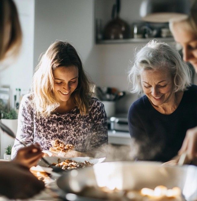 A smiling young woman and an older woman serving food in a sunlit kitchen, representing the joy of social connection and relief from anxiety