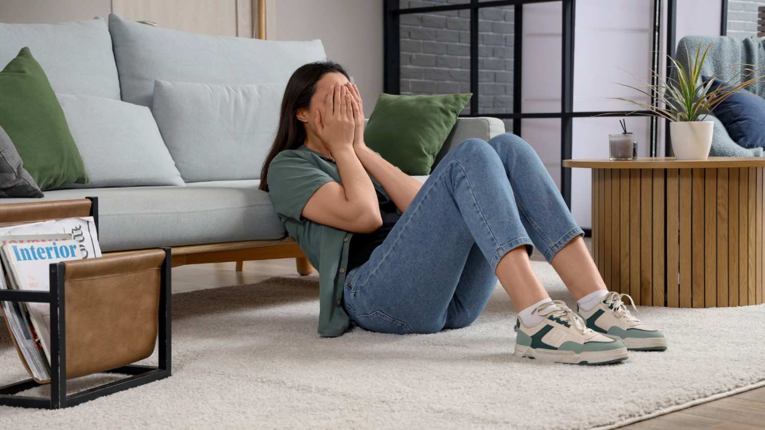Woman Sitting on Living Room Floor Feeling Stressed at Home A woman sitting on the carpeted floor beside a sofa with her hands covering her face, appearing stressed or overwhelmed in a modern living room.